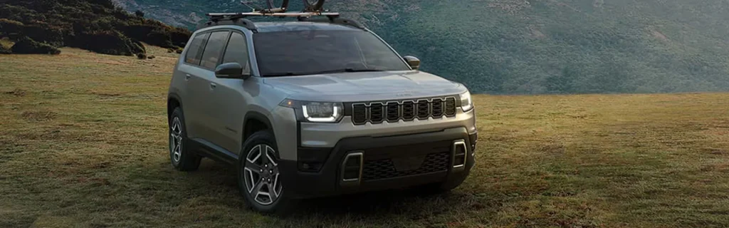 Silver Jeep SUV with a roof-mounted bike rack parked on a grassy field against a scenic mountain backdrop.